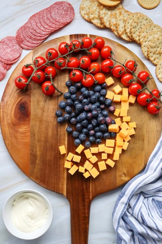 Blueberries placed in the center of a wooden board with cherry tomatoes arranged along the top edge and cheddar cubes to the side, showing the base layout for assembling a patriotic meat and cheese platter.