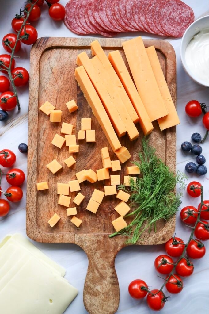 Cheddar cheese cut into cubes and sticks on a wooden board, with cherry tomatoes, salami, and fresh herbs nearby, showing a prep step for assembling a patriotic meat and cheese platter.