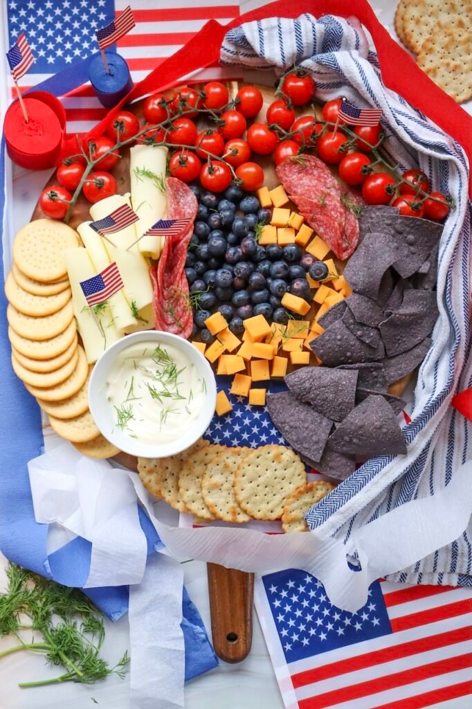 A finished 4th of July charcuterie board on a wooden platter with blueberries in the center, cherry tomatoes on the vine, cheddar cubes, Swiss cheese slices, crackers, blue tortilla chips, salami, and a bowl of dip, styled with small American flags for a festive presentation.