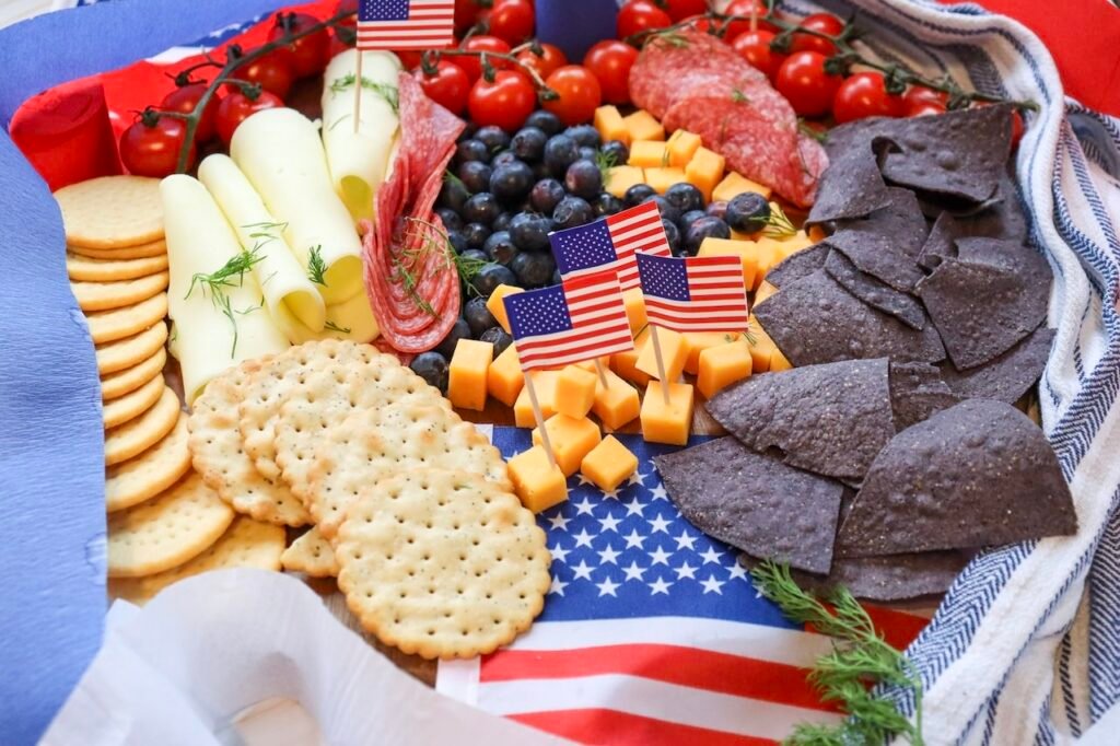 Angled view of a 4th of July charcuterie board with crackers, rolled cheese slices, blueberries, cherry tomatoes on the vine, cheddar cubes with small American flags, salami, and blue tortilla chips arranged on a wooden board.