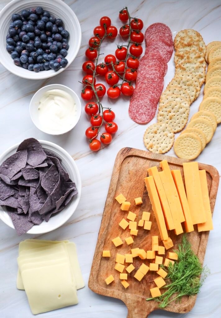 Ingredients for a 4th of July charcuterie board laid out on a countertop, including blueberries, cherry tomatoes on the vine, salami slices, crackers, blue tortilla chips, Swiss cheese, cheddar cubes and sticks on a wooden board, and a small bowl of dip.