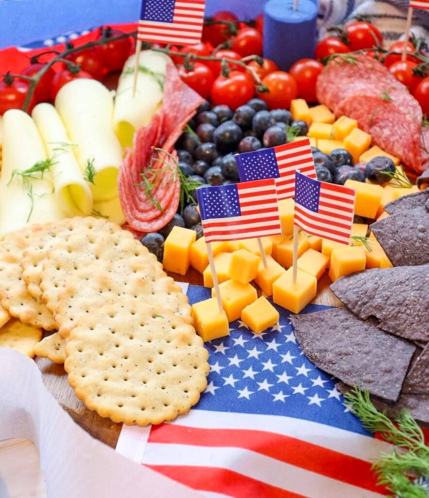 Close-up of a 4th of July charcuterie board with crackers, cheddar cheese cubes topped with small American flags, blueberries, salami, rolled cheese slices, and blue tortilla chips arranged on a wooden board.