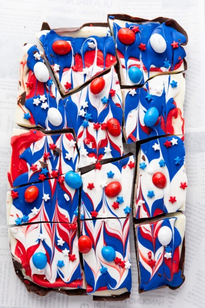 Broken pieces of red, white, and blue treat topped with sprinkles and candy-coated chocolates on parchment paper.