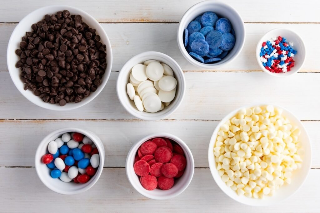 Overhead view of small bowls filled with white chocolate chips, semi-sweet chocolate chips, red and blue candy melts, white candy melts, patriotic sprinkles, and red white and blue candies arranged on a white wooden surface for making chocolate bark.