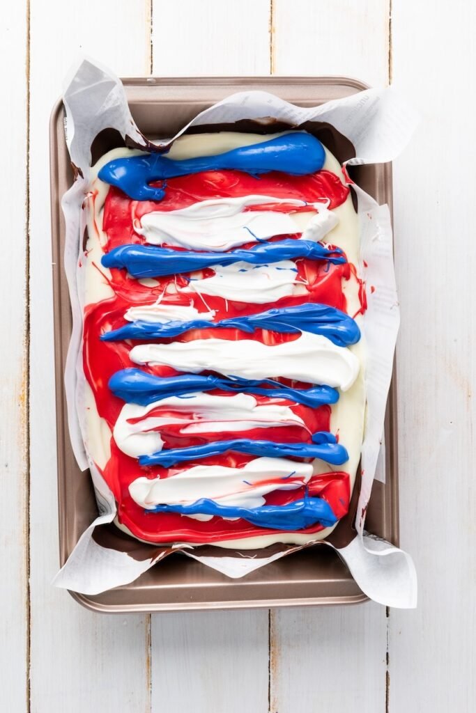 Red, white, and blue candy melts drizzled in thick stripes over layered chocolate on a parchment-lined tray before swirling.