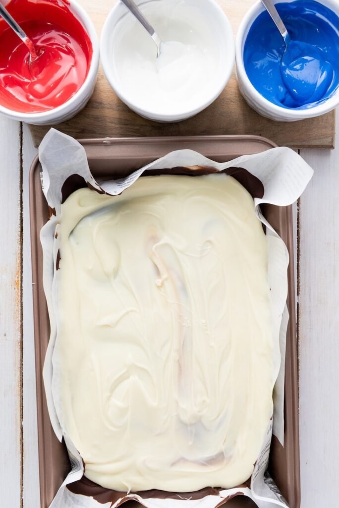 Smooth melted white chocolate spread evenly over a layer of semi-sweet chocolate on a parchment-lined pan, showing the second layer of 4th of July chocolate bark.