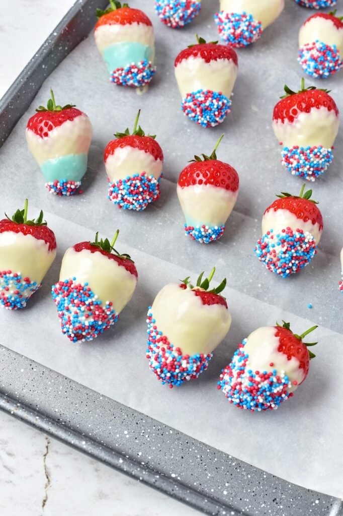Finished chocolate covered strawberries decorated with red, white, and blue sprinkles arranged on a parchment-lined tray for a patriotic dessert.