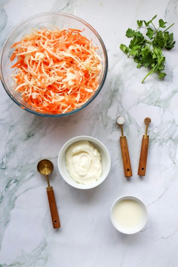 Overhead view of shredded cabbage and carrots in bowls with mayonnaise, vinegar, parsley, and seasonings arranged on a marble surface for making coleslaw.