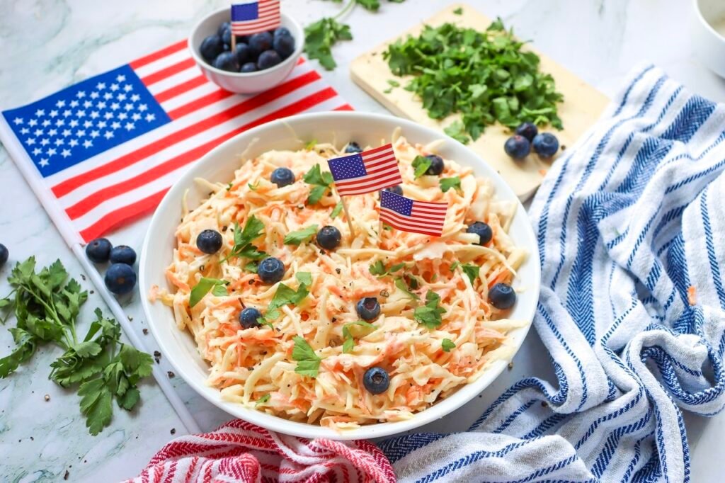 Overhead view of a bowl of creamy coleslaw with shredded cabbage and carrots, topped with blueberries and herbs, surrounded by an American flag and kitchen linens for a 4th of July setting.