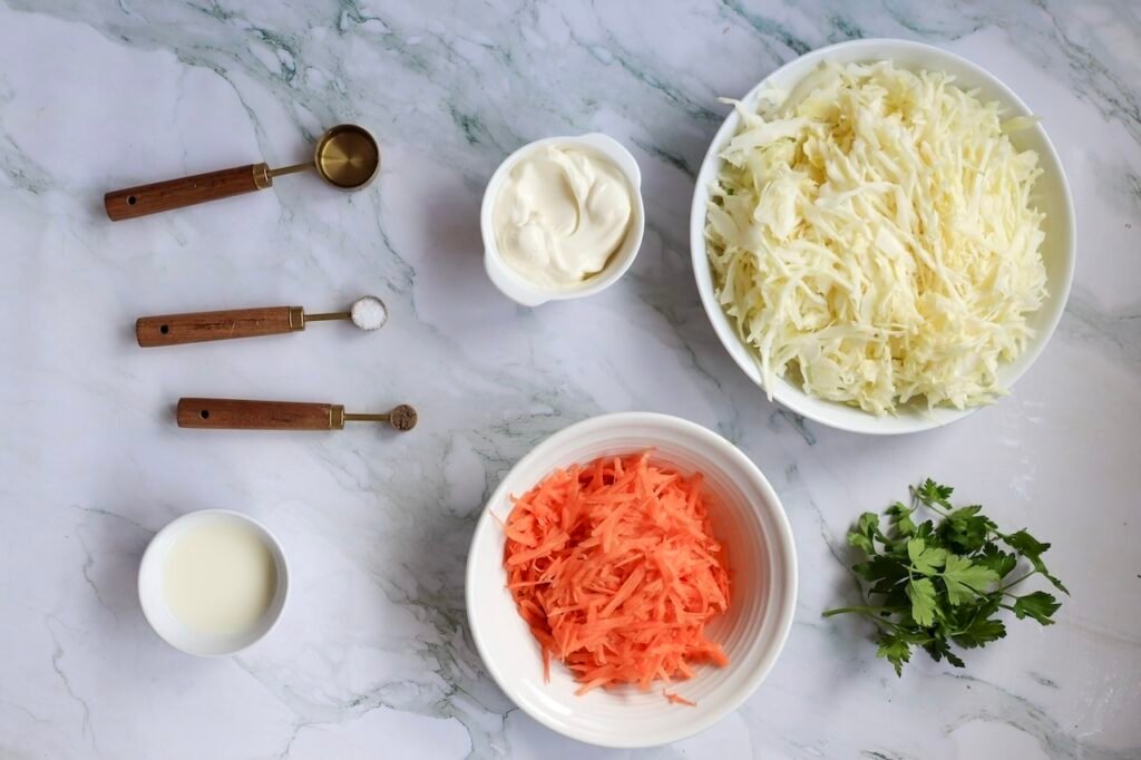 Overhead view of shredded cabbage and carrots in bowls with mayonnaise, vinegar, parsley, and seasonings arranged on a marble surface for making coleslaw.
