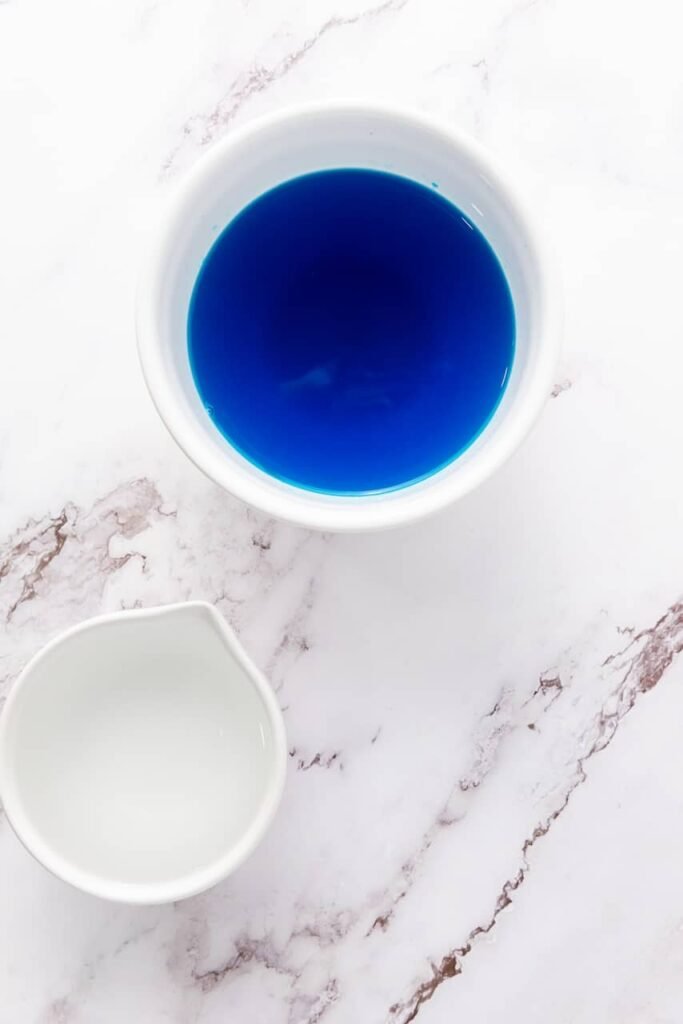 Overhead view of fully mixed blue gelatin liquid in a bowl with water nearby, showing the prepared blue layer for 4th of July jello shots.