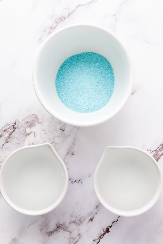Overhead view of blue gelatin powder in a bowl with two small pitchers of water, showing ingredients for the blue layer of 4th of July jello shots.