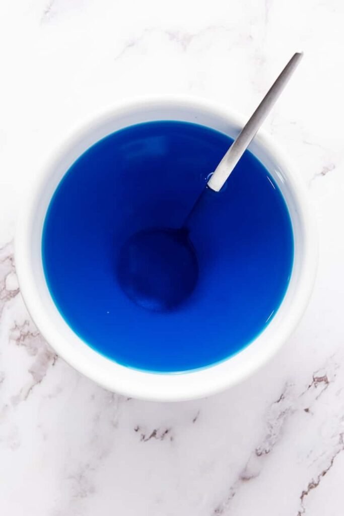 Overhead view of blue gelatin being stirred with a spoon in a bowl, showing the mixing step for 4th of July jello shots.