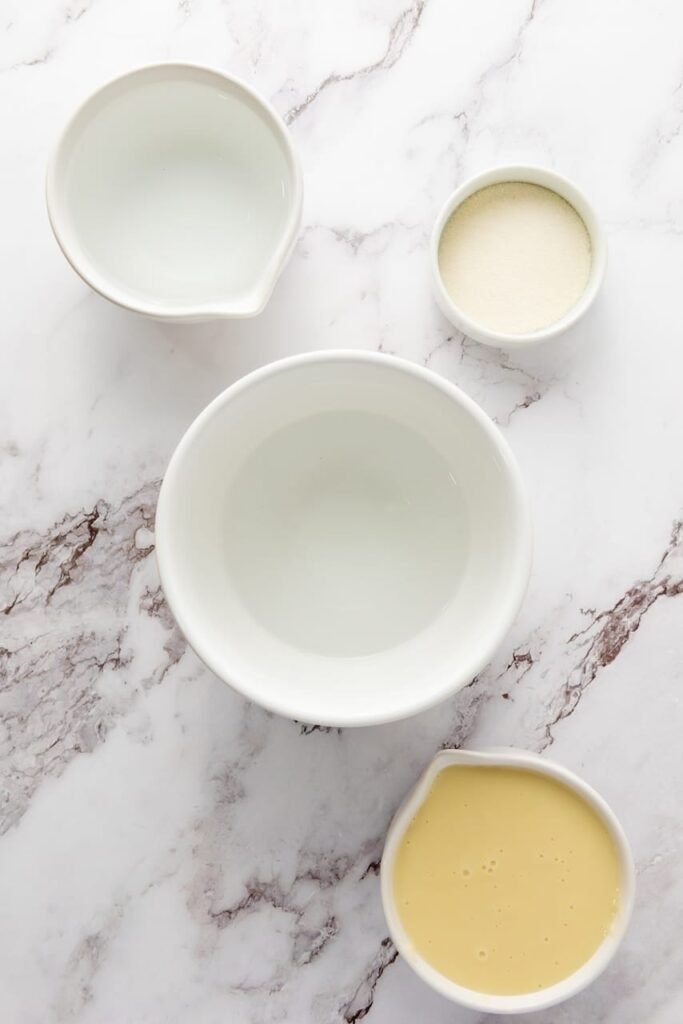 Overhead view of ingredients for 4th of July jello shots including water, gelatin powder, and sweetened condensed milk in white bowls on a marble surface