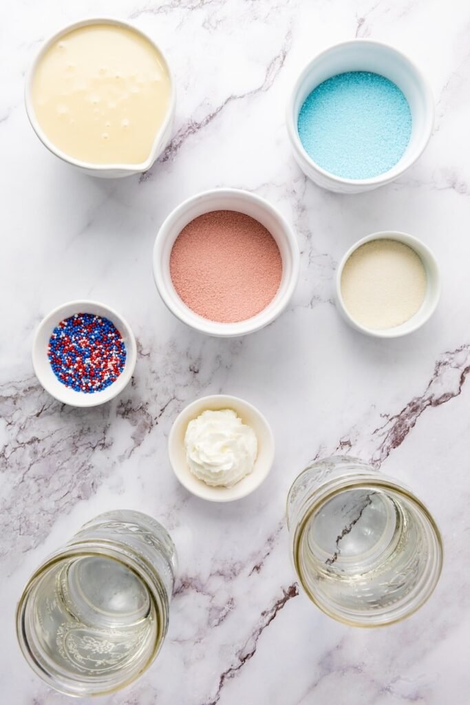 Overhead view of ingredients for 4th of July jello shots including blue and red gelatin powder, unflavored gelatin, sweetened condensed milk, whipped cream, sprinkles, and water on a marble surface.