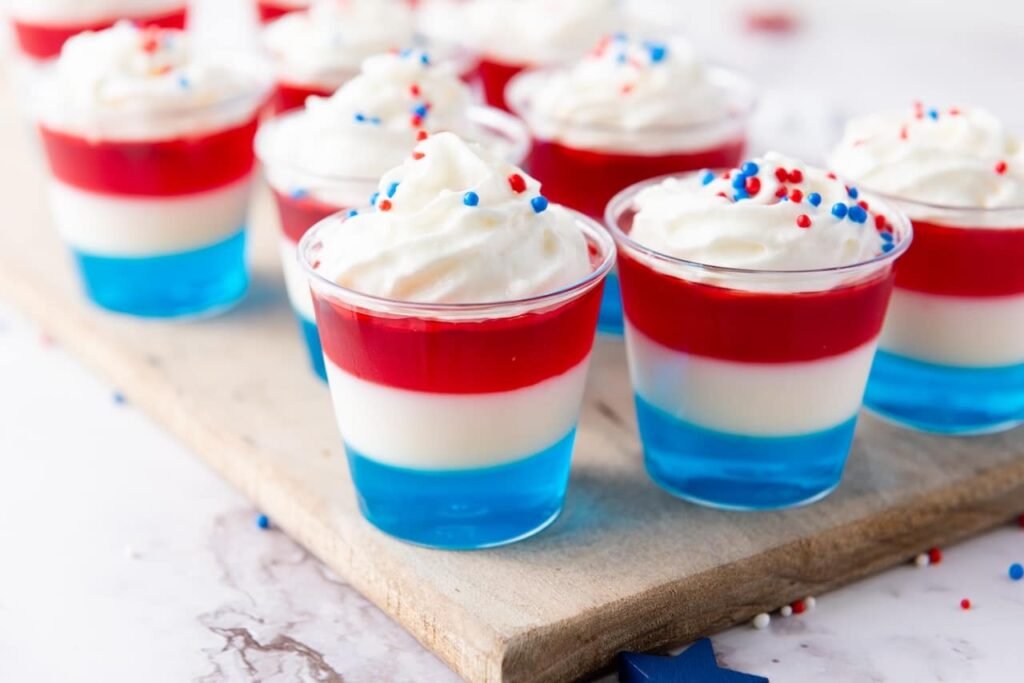 Side view of layered 4th of July jello shots with blue, white, and red layers topped with whipped cream and sprinkles on a wooden board.