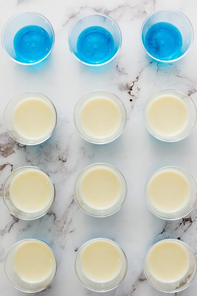 Overhead view of 4th of July jello shots in plastic cups showing blue gelatin base with creamy white layer being added in rows on a marble surface
