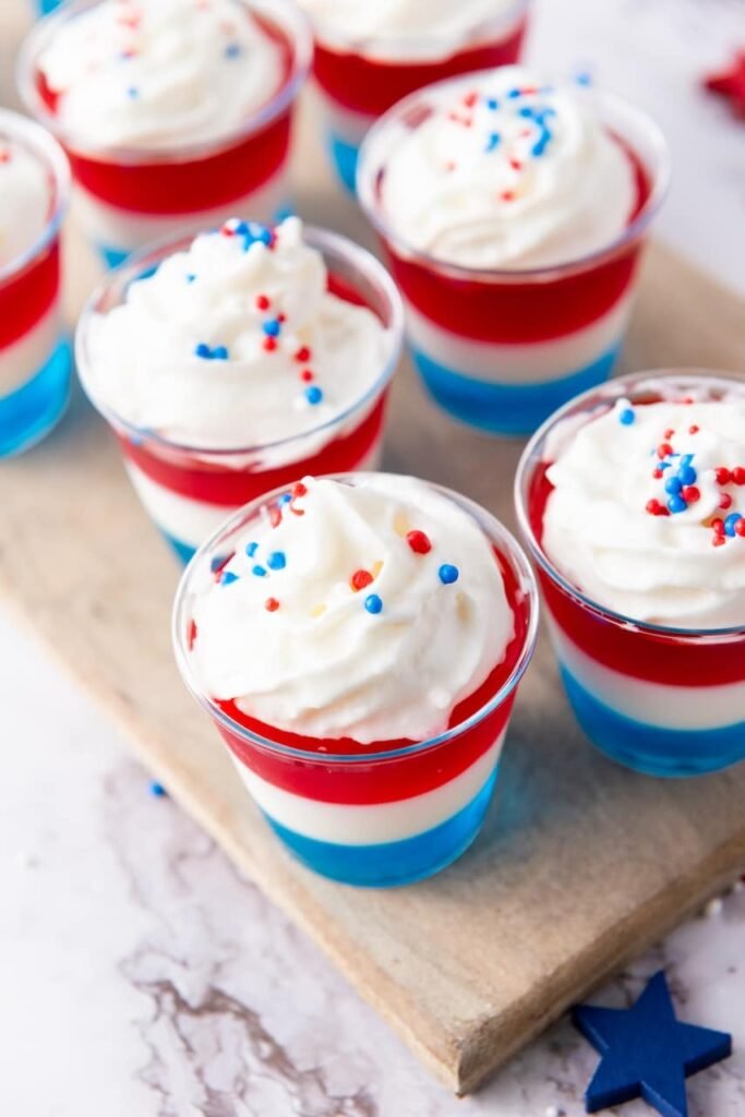 Close-up of layered 4th of July jello shots with red white and blue layers topped with whipped cream and festive sprinkles in clear cups