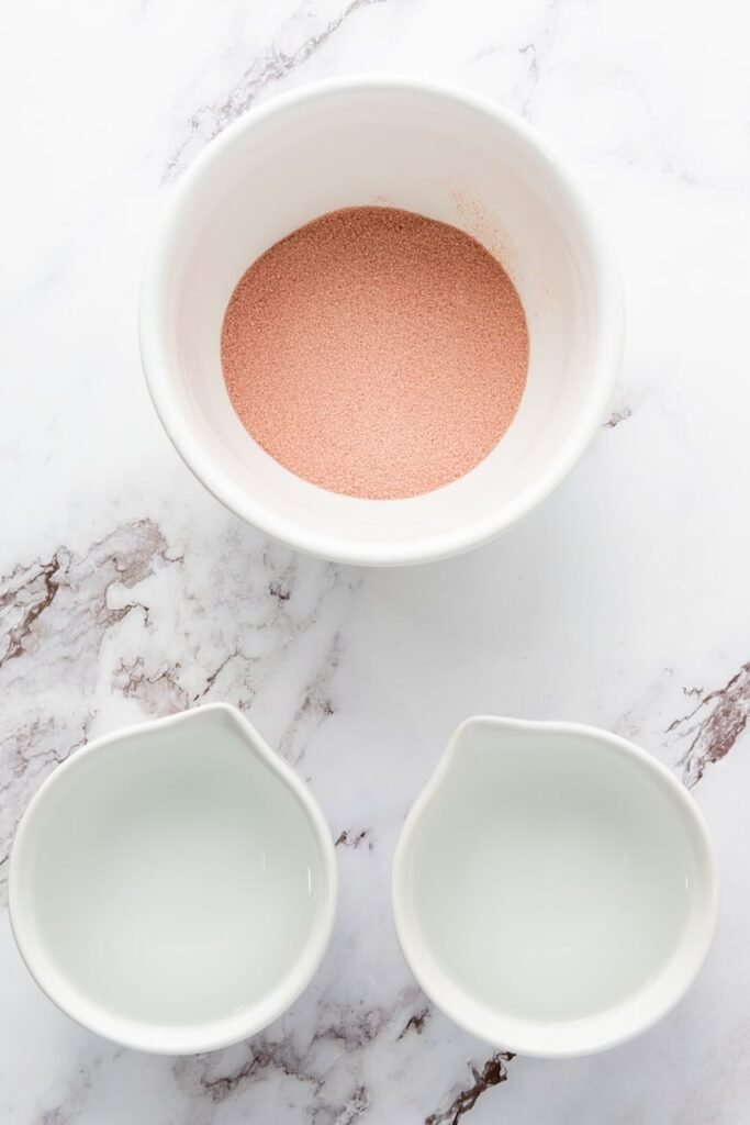 Overhead view of red gelatin powder in a bowl with two small pitchers of water, showing ingredients for the red layer.