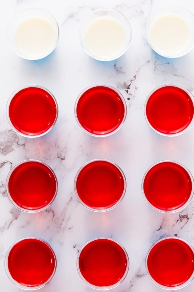 Overhead view of red gelatin layer set in small plastic cups with white layer above, showing layered in progress.
