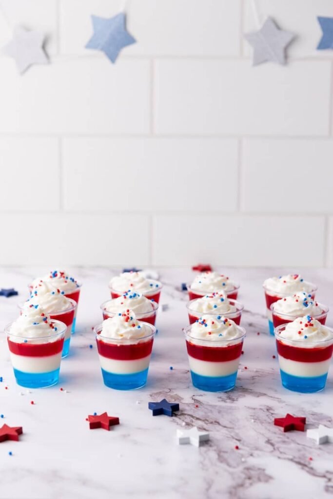 Red white and blue 4th of July jello shots layered in clear cups topped with whipped cream and patriotic sprinkles on a marble surface