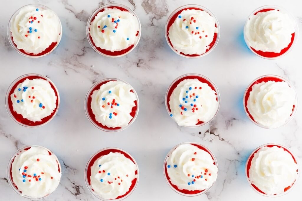 Overhead view of 4th of July jello shots topped with whipped cream and red, white, and blue sprinkles in small cups.