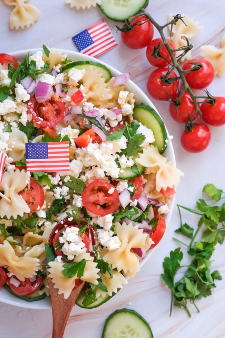 Close-up view of 4th of July pasta salad with bowtie pasta, cherry tomatoes, cucumber slices, red onion, feta cheese, and parsley, decorated with small American flags on a white surface.