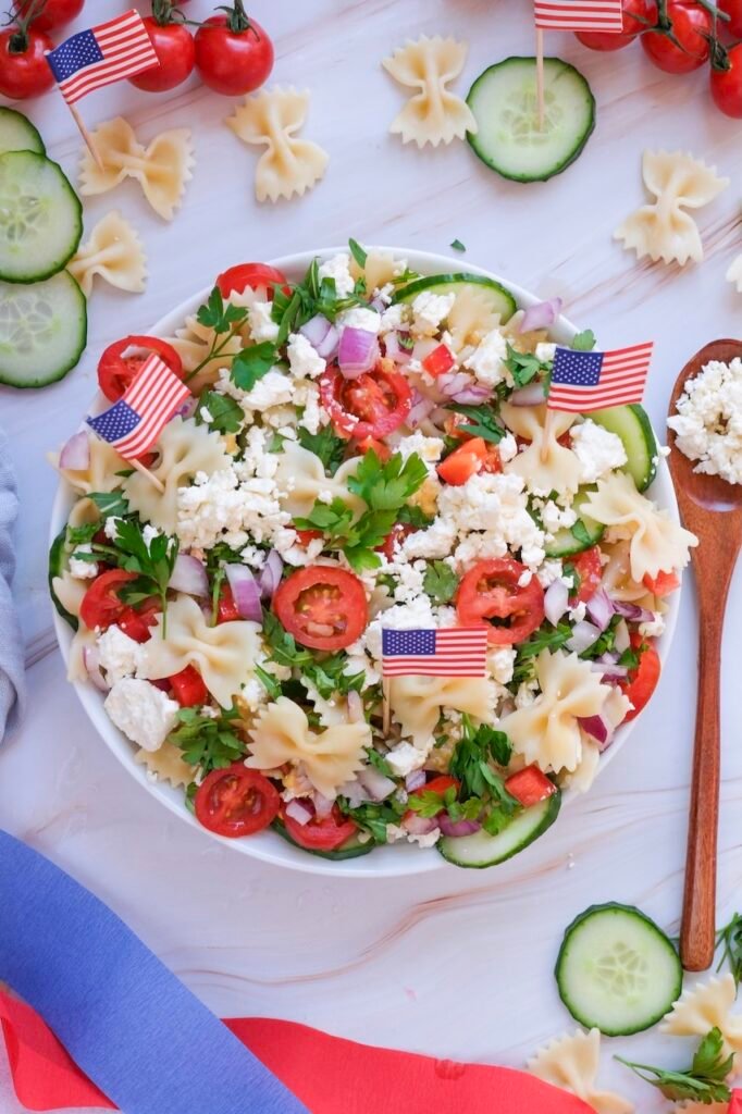 Overhead view of a bowl with bowtie pasta, cherry tomatoes, cucumber, red onion, feta cheese, and parsley, decorated with small American flags on a light surface.