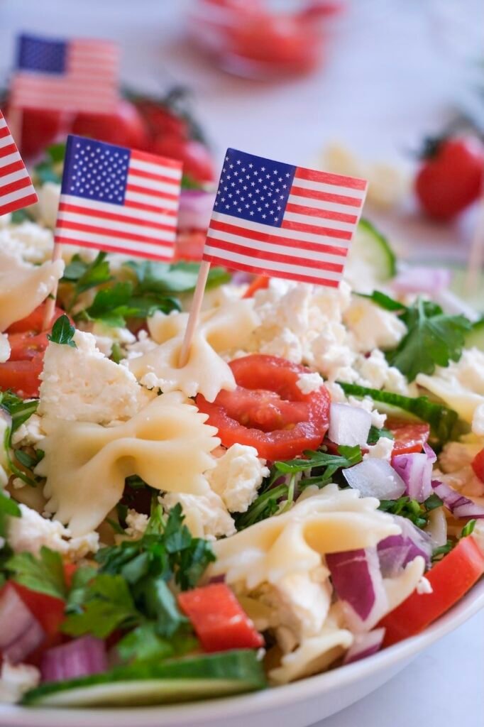Close-up of 4th of July pasta salad with bowtie pasta, cherry tomatoes, cucumber, red onion, feta cheese, and parsley, topped with small American flags in a festive presentation.