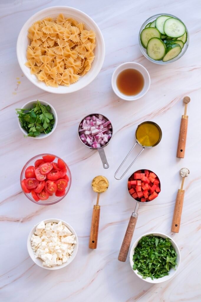 Overhead view of ingredients for 4th of July pasta salad including cooked bowtie pasta, cherry tomatoes, cucumber slices, red bell pepper, red onion, arugula, feta cheese, parsley, and a small bowl of vinaigrette dressing arranged on a light surface.