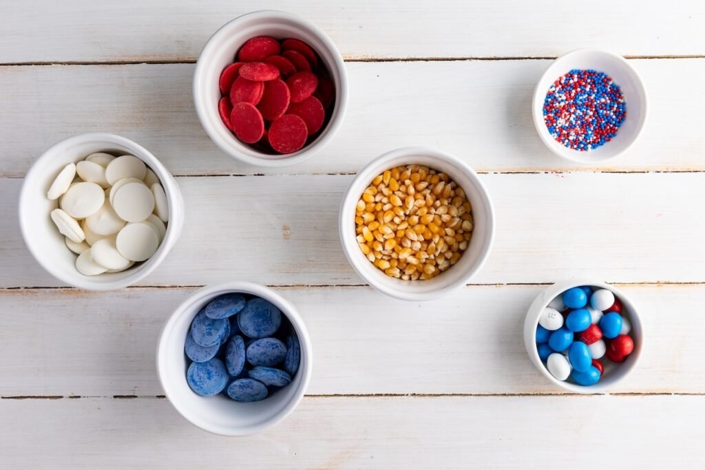 Six small white bowls arranged on a white wooden surface, each holding ingredients for 4th of July popcorn, including popcorn kernels, red and blue candy melts, white chocolate wafers, patriotic sprinkles, and red, white, and blue M&M candies.