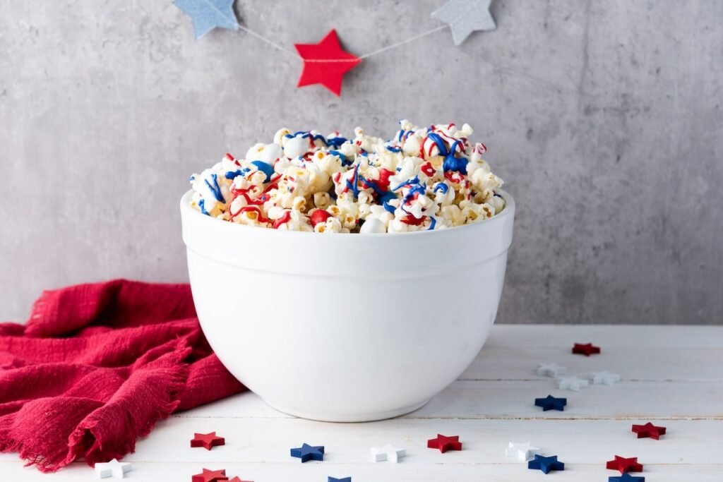 A large white bowl filled with popcorn drizzled in red, white, and blue candy melts, decorated with sprinkles and M&Ms, set on a table with patriotic decorations and a red cloth in the background.