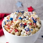 A white bowl filled with popcorn drizzled in red, white, and blue candy melts and topped with M&M candies and sprinkles, set on a table with patriotic decorations in the background.