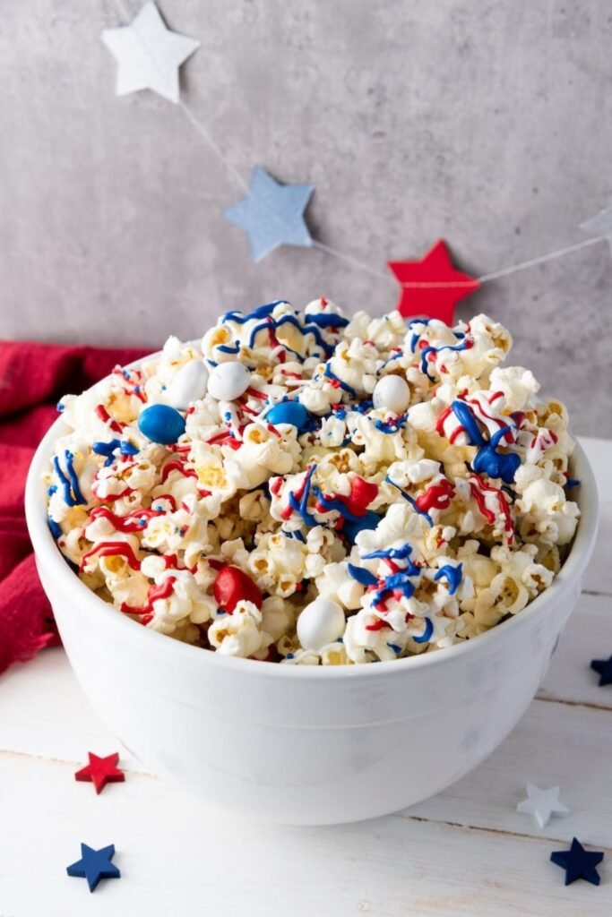 A white bowl filled with popcorn drizzled in red, white, and blue candy melts and topped with M&M candies and sprinkles, set on a table with patriotic decorations in the background.