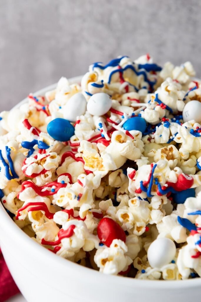 Close-up view of a bowl of popcorn topped with red, white, and blue candy drizzle, M&M candies, and sprinkles, showing texture and detail of the finished 4th of July popcorn.