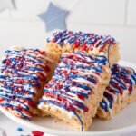 Stack of 4th of July rice krispie treats on a white cake stand, decorated with red, white, and blue candy drizzle and sprinkles.
