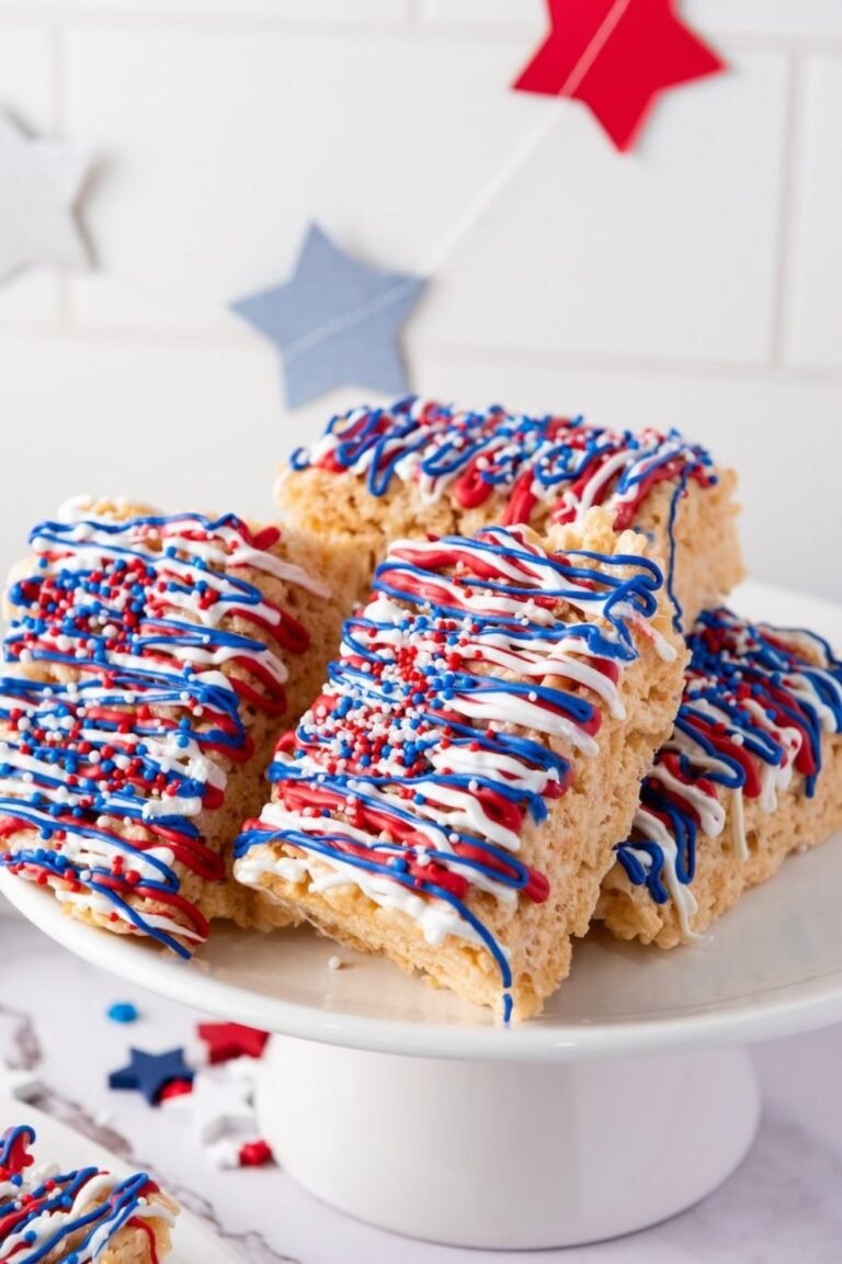 Stack of 4th of July rice krispie treats on a white cake stand, decorated with red, white, and blue candy drizzle and sprinkles.