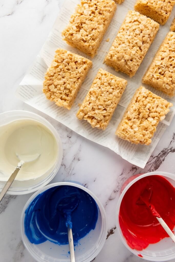 Overhead view of easy july 4th desserts cut into rectangles with bowls of melted red, white, and blue candy melts ready for dipping.
