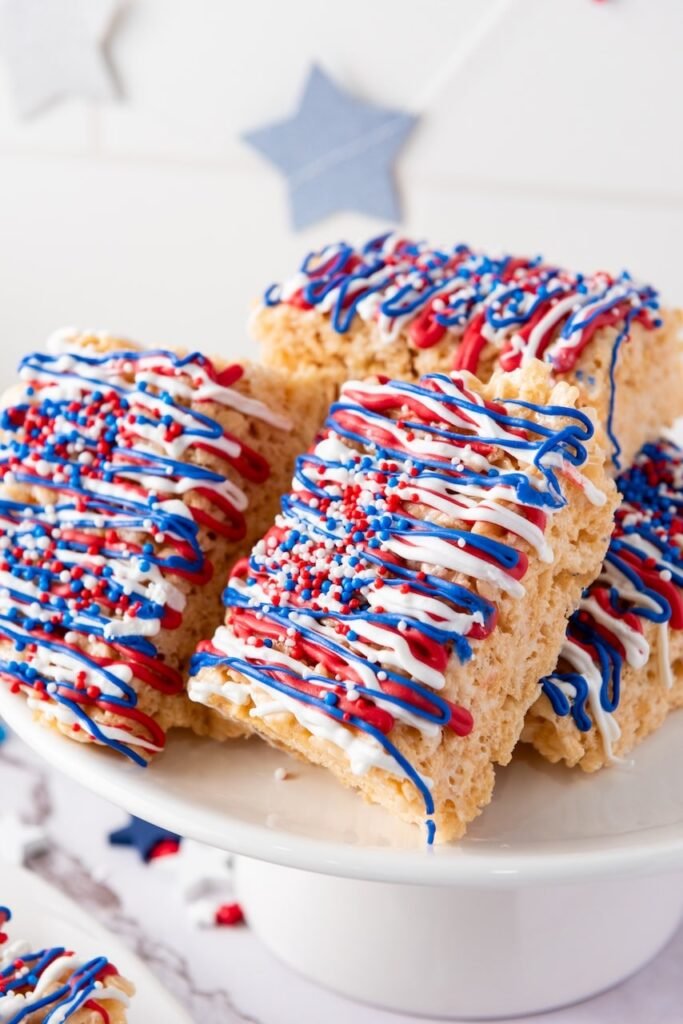 Close-up of 4th of July rice krispie treats stacked on a cake stand, decorated with red, white, and blue candy drizzle and sprinkles.