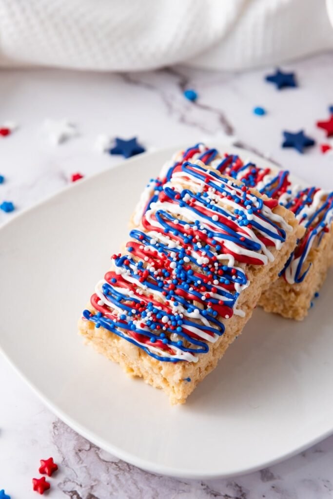Close-up of 4th of July rice krispie treats on a plate, decorated with red, white, and blue candy drizzle and sprinkles.