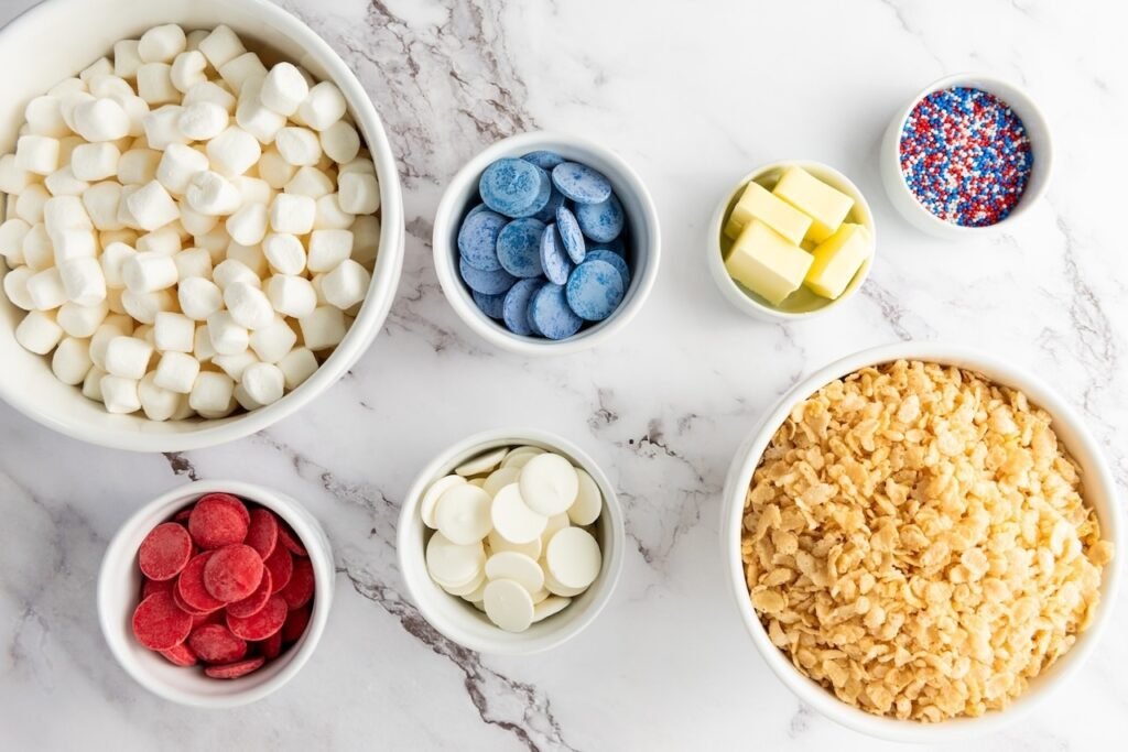 Overhead view of ingredients for 4th of July Rice Krispie treats, including cereal, mini marshmallows, butter, red white and blue candy melts, and patriotic sprinkles arranged in bowls.