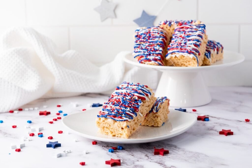 4th of July rice krispie treats served on plates and a cake stand, decorated with red, white, and blue drizzle and sprinkles in a festive setting.