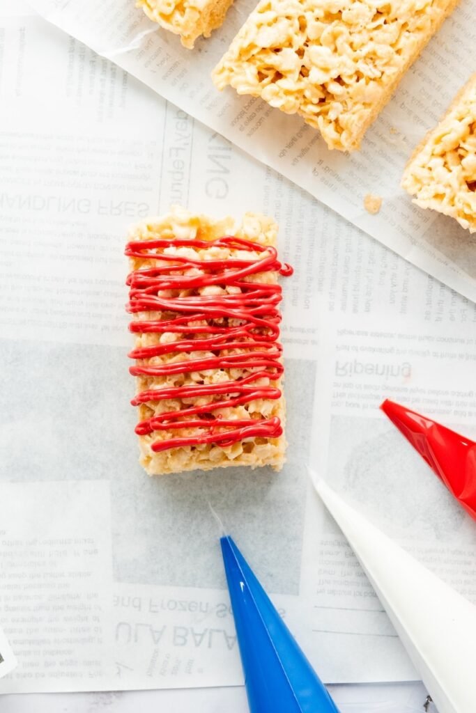 Close-up of a 4th of July rice krispie treat rectangle with red candy melt drizzle on top, with piping bags nearby for decorating.