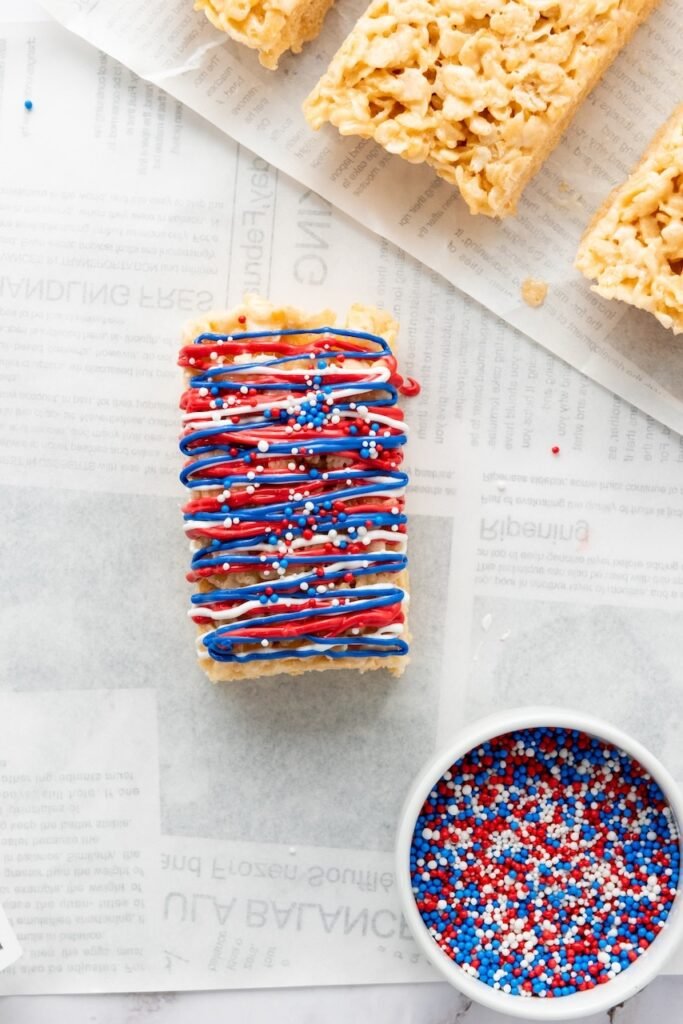 Close-up of a 4th of July rice krispie treat decorated with red, white, and blue candy drizzle and sprinkles, with a bowl of patriotic sprinkles nearby.