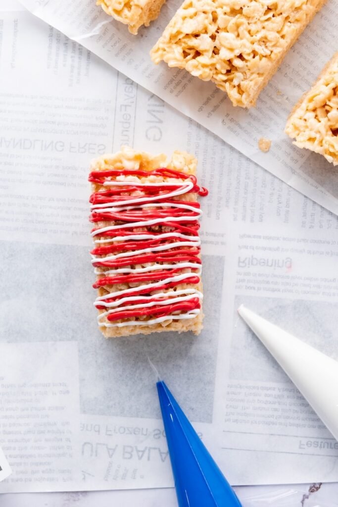 Close-up of a 4th of July rice krispie treat with red and white candy melt drizzle on top, with piping bags nearby for decorating.