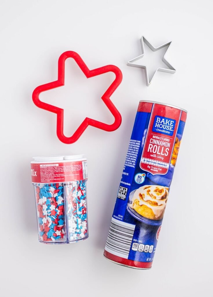 Overhead view of ingredients for star-shaped cinnamon rolls including a can of cinnamon rolls, red and silver star cookie cutters, and red, white, and blue sprinkles on a light background.