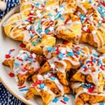 Plate of star-shaped cinnamon rolls drizzled with icing and topped with red, white, and blue sprinkles, set on a festive patriotic background.