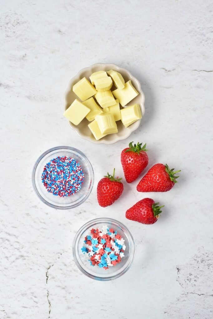 Fresh strawberries, white chocolate pieces, and red, white, and blue sprinkles arranged in small bowls on a light surface as ingredients for 4th of july strawberries.