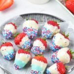 Chocolate covered strawberries decorated with red, white, and blue sprinkles arranged on a serving plate with parchment paper for a patriotic dessert display.