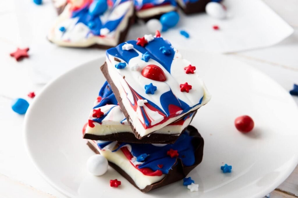 Stacked pieces of red, white, and blue chocolate bark with sprinkles and candy on a white plate, showing the finished no-bake 4th of July dessert.